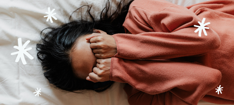 Woman with black hair wearing an orange sweater lying on a white bed with her hands over her eyes