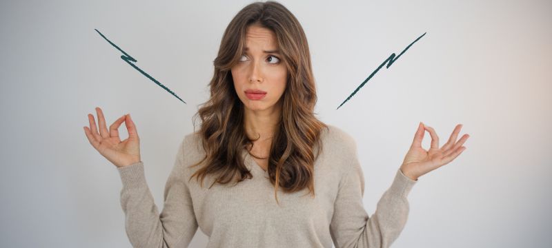 White woman with long brown hair in a pale jumper holds her hands up in a traditional meditation pose while looking confused. Two spiky illustrated lines sit above her shoulders