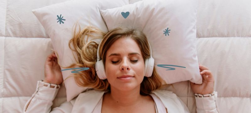 Woman lying in bed with hands under pillow, eyes closed & wearing headphones